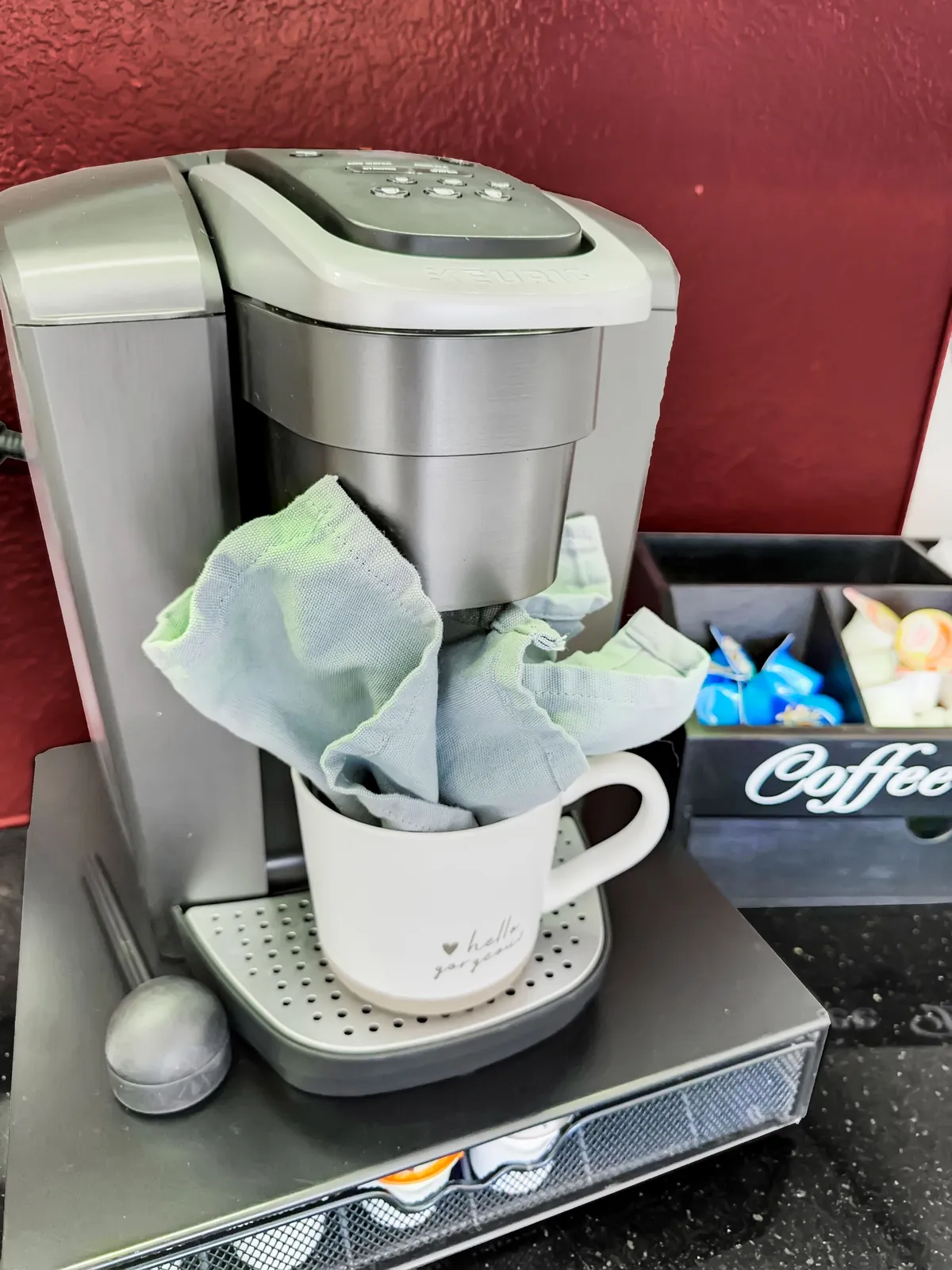 A close-up of a coffee maker set on a countertop, accompanied by a mug on a cloth and a drawer filled with coffee pods. The cozy vignette suggests the simple pleasure of brewing coffee at home before heading out or settling in to work.