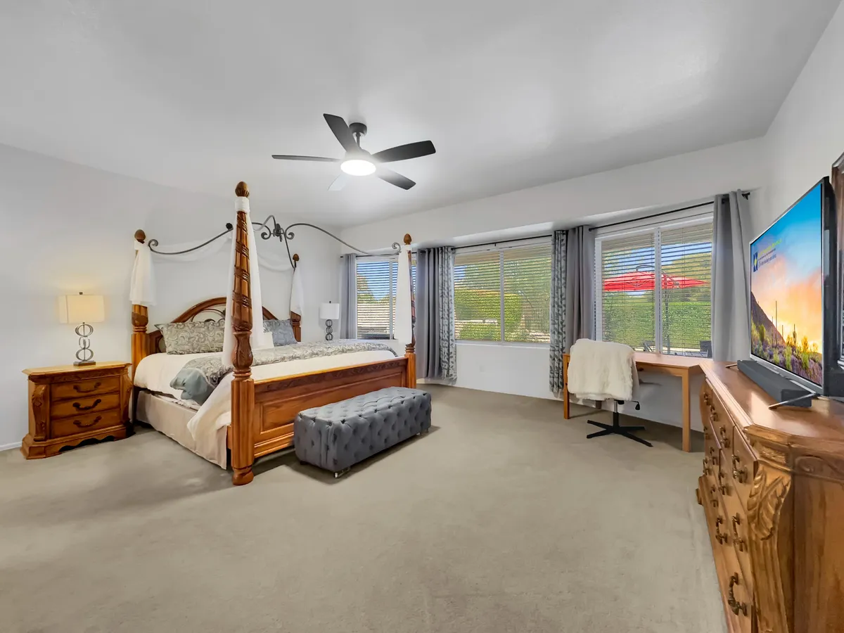 A spacious bedroom featuring a classic four-poster bed with crisp linens, a nightstand with a lamp, chest of drawers, large windows letting in ample natural light, and a desk with a chair in the corner. The design is calm and inviting, with soft colors and uncluttered decor.