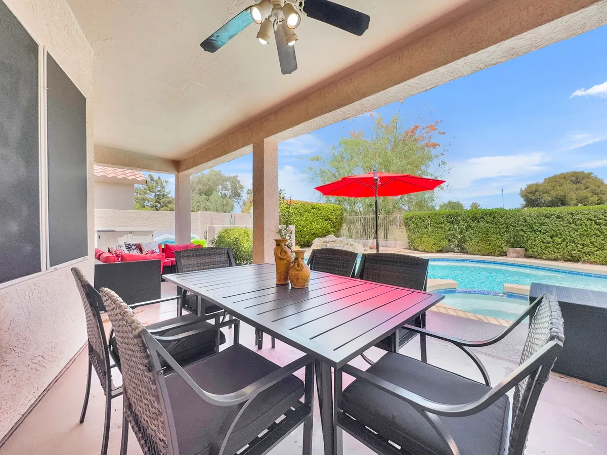 A sun-drenched backyard scene featuring a spacious dining table set beside a sparkling swimming pool. Red umbrellas provide stylish shade over the lounging and dining areas, while lush landscaping completes the inviting, social atmosphere. Ample outdoor seating suggests plenty of room for group gatherings, outdoor meals, or poolside celebrations.