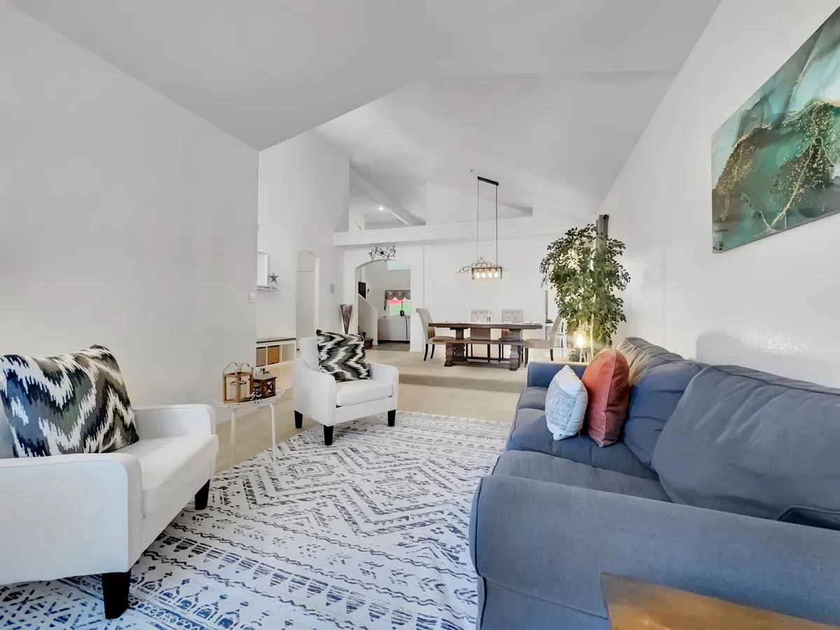 A warmly lit open-plan living and dining area featuring a comfortable sofa and armchairs adorned with decorative pillows, an indoor plant adding a touch of greenery, and a dining table set for family or group meals. Large windows allow sunlight to stream in, highlighting the welcoming, inviting atmosphere.