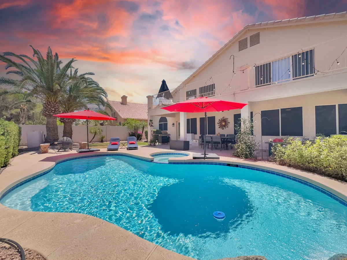 An inviting outdoor pool area surrounded by palm trees and red umbrellas, with a spacious deck for lounging and social gatherings. The setting is perfect for relaxing swims or evening gatherings, and the pool is highlighted under the warm glow of sunset and string lights.