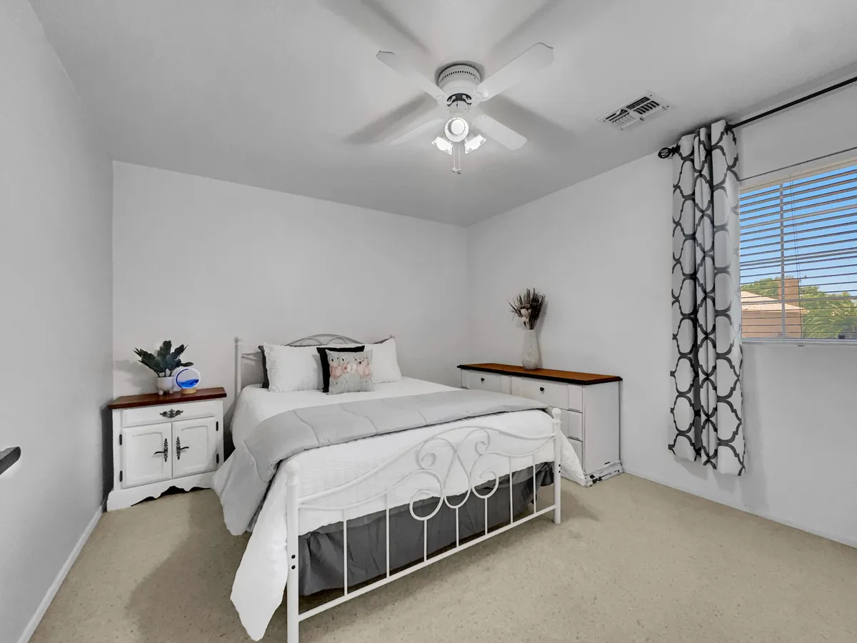 A cozy bedroom with a bed dressed in white linens and decorative pillows, a nightstand topped with a plant, draped curtains, and a window with blinds that let in natural light. The atmosphere is peaceful and restful, ideal for unwinding after a busy day.