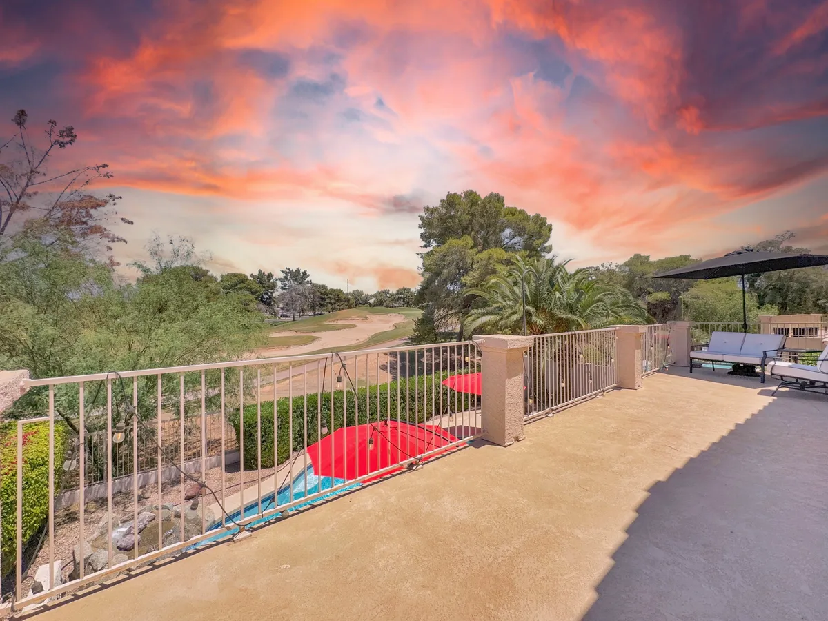 A tranquil balcony scene overlooking the pool area, complete with red umbrellas and lush landscape, offering expansive views towards the surrounding scenery and glimpses of the golf course. The setup invites leisurely lounging and captures the essence of enjoying sunsets or quiet moments after active exploration.