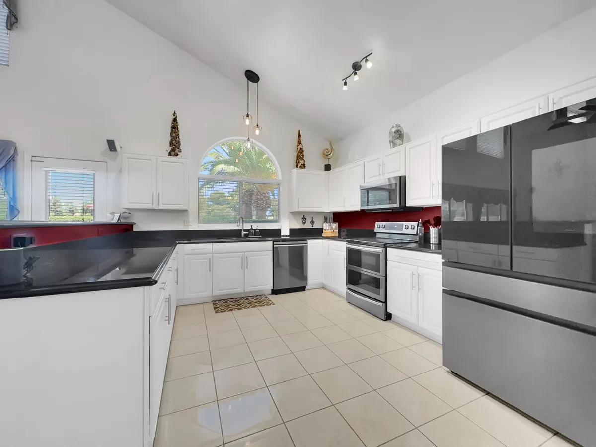 A modern, cheerful kitchen outfitted with black countertops, stainless steel appliances, and white cabinetry. A large window offers a view of palm trees outside, bathing the kitchen in natural light and connecting the interior with the landscape beyond.