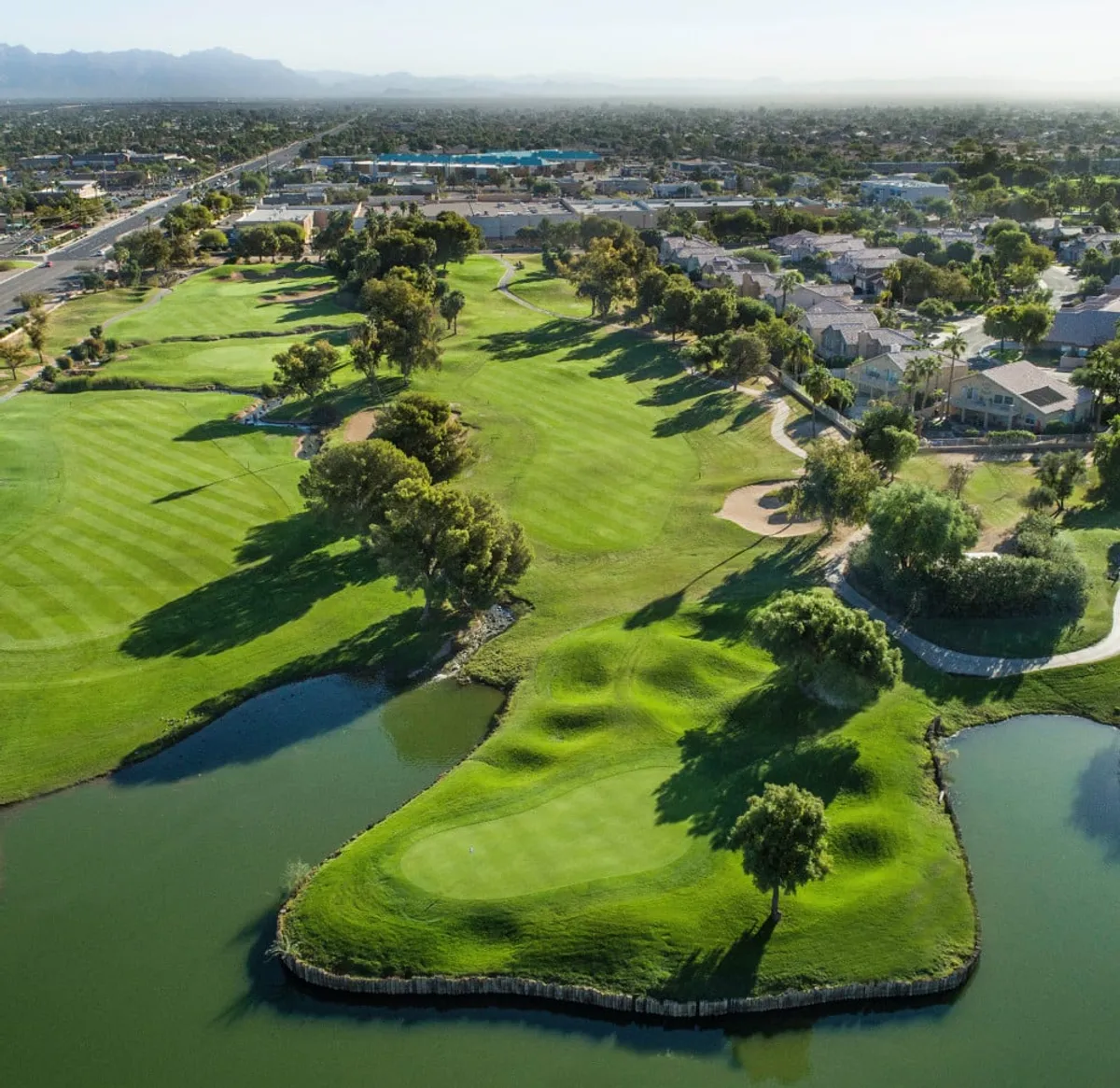 This image captures a panoramic view of the Superstition Springs Golf Club, with lush green fairways, sparkling water features, and surrounding residential areas. Golden hour lighting bathes the scene, accentuating the vibrant landscape and inviting viewers into the relaxing, recreational mood of the golf course environment.