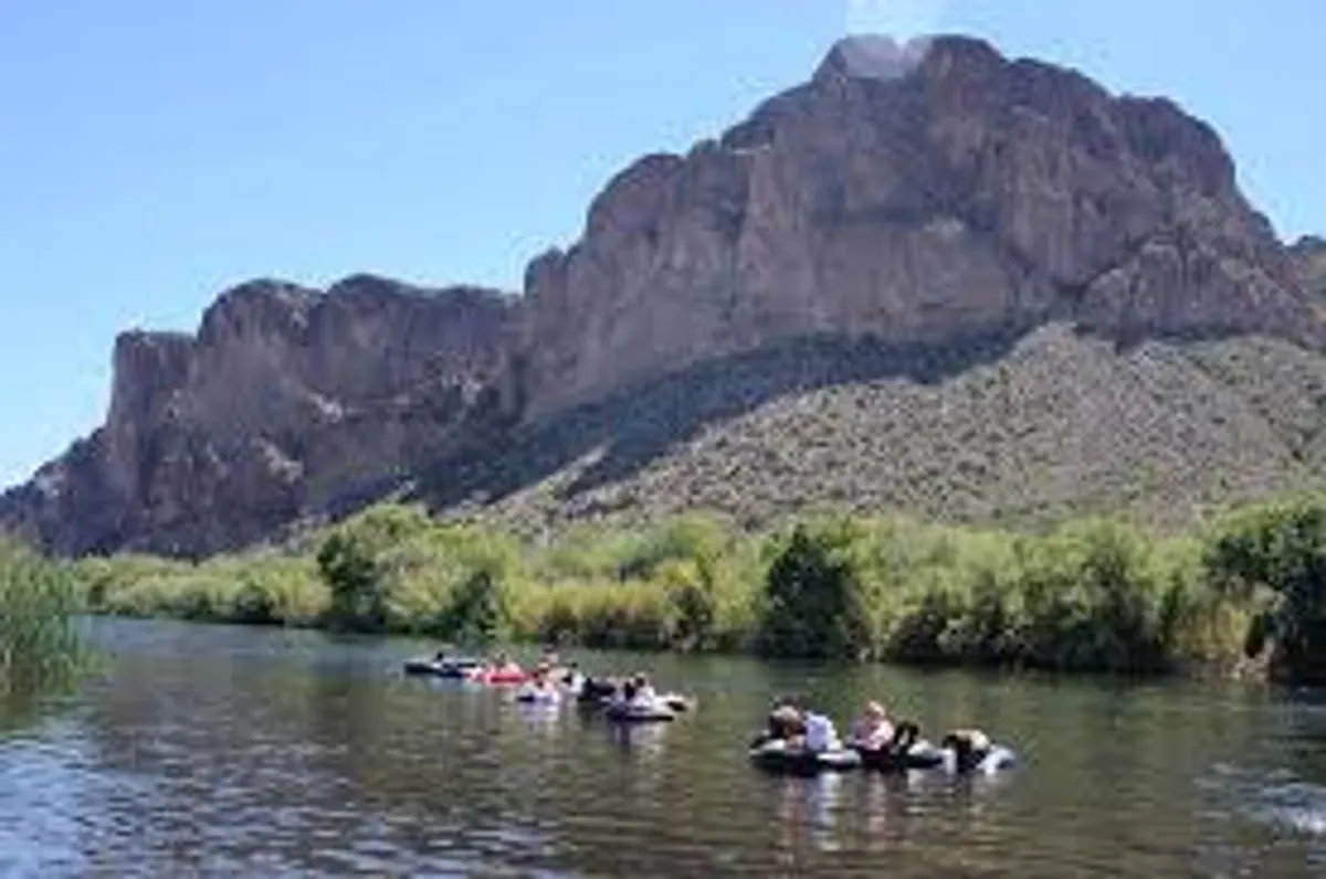 A beautiful outdoor scene featuring a mountainous backdrop with a flowing river in the foreground, where people are seen tubing and enjoying the water. The landscape is lush and natural, evoking a sense of adventure and outdoor recreation.